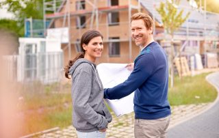 junges Paar mit Bauplan in der Hand vor Haus-Baustelle