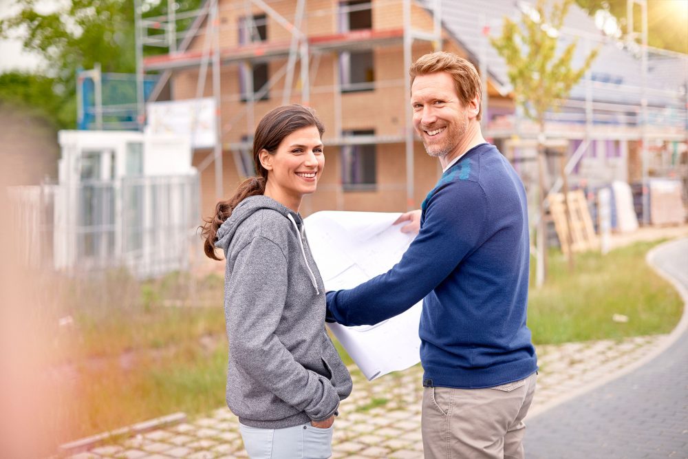 junges Paar mit Bauplan in der Hand vor Haus-Baustelle