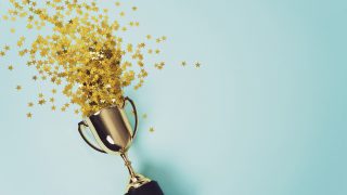 Goldener Pokal mit zwei Griffen, aus dem viele kleine goldene Sterne nach oben fliegen, vor hellblauem Hintergrund.