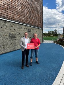 Steffen Burdack von der Rheinhessen Sparkasse und Rainer Uhrig vom JFV Bürstadt halten bei der Spendenübergabe für den Bildungs- und Sportcampus in Bürstadt ein rotes Schild mit dem Logo der Rheinhessen Sparkasse.