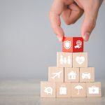 Eine Hand legt einen roten Baustein auf die Spitze einer Pyramide aus naturbelassenen Bausteinen. Auf den Bausteinen sind Piktogramme erkennbar.