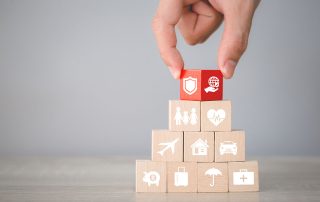 Eine Hand legt einen roten Baustein auf die Spitze einer Pyramide aus naturbelassenen Bausteinen. Auf den Bausteinen sind Piktogramme erkennbar.