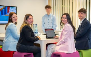Auszubildende der Rheinhessen Sparkasse sitzen gemeinsam am Meeting Point und schauen in die Kamera.