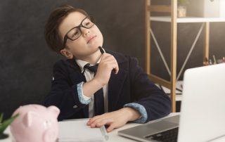 Ein Junge mit Anzug und Krawatte sitzt vor einem Laptop, daneben ein Sparschwein. Mit einem Stift kratzt er sich am Kinn.