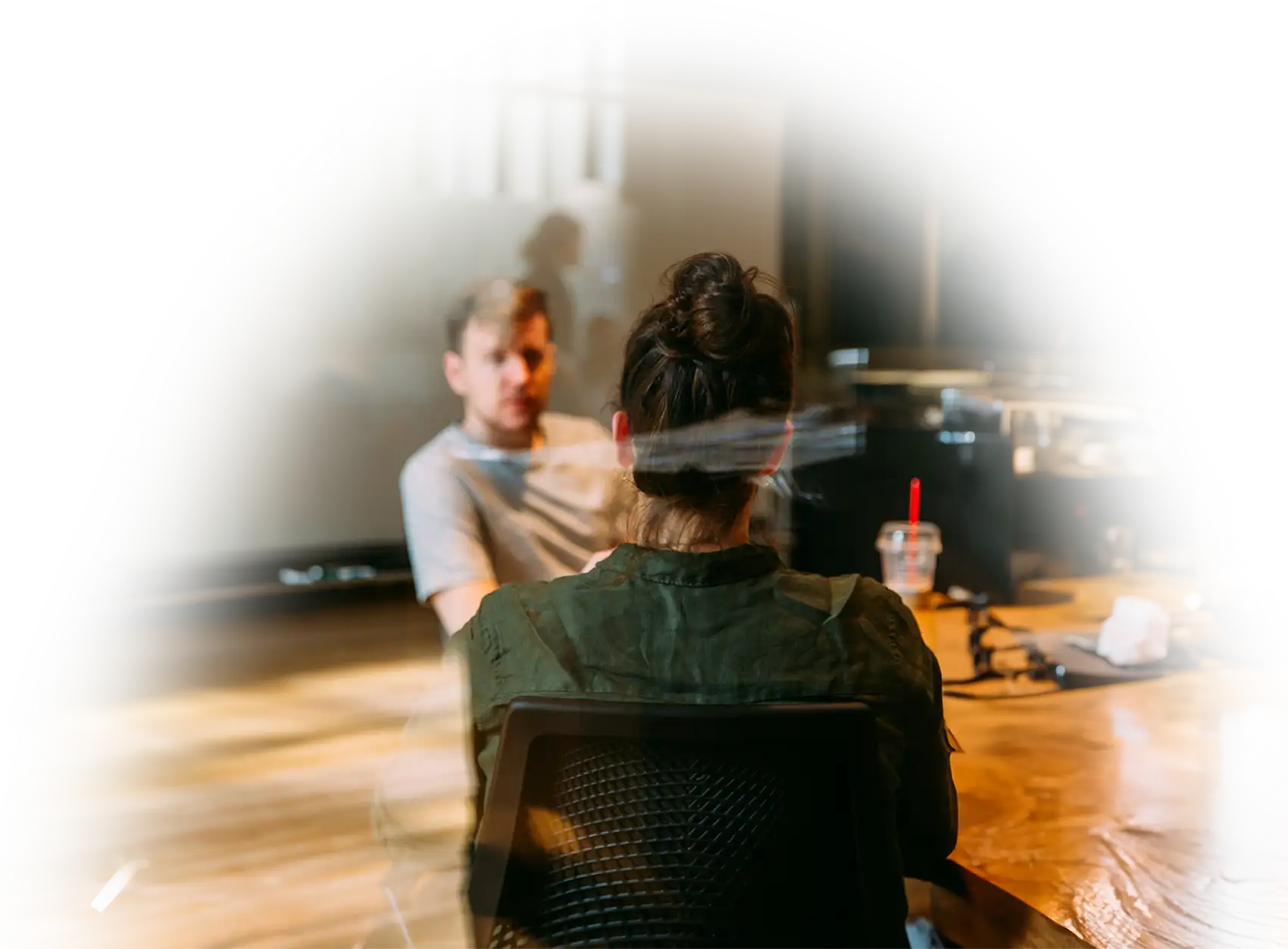 Zwei Personen sitzen sich an einem Tisch gegenüber und unterhalten sich, im Hintergrund ist ein Büro oder Besprechungsraum zu erkennen.